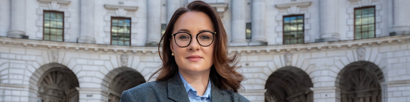 Yulia Svyrydenko wearing a grey blazer and light blue shirt stands in front of a grand, historic architectural structure. The setting appears to be outdoors, with stone arches and windows visible in the background. The overall mood is professional and urban, with neutral tones dominating the palette. No visible text or numbers are present in the image.