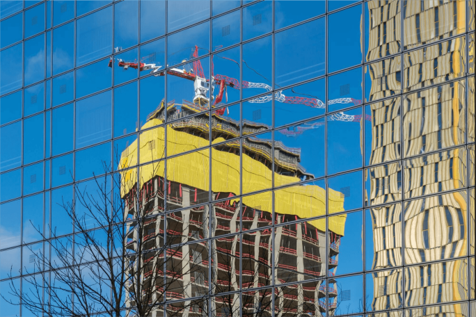 A high-rise building under construction is reflected in the glass windows of a nearby structure. The construction site features a prominent yellow covering and a visible crane against a clear blue sky. The reflection creates a distorted, abstract effect, with tree branches also visible in the foreground. No visible text or numbers are present in the image.
