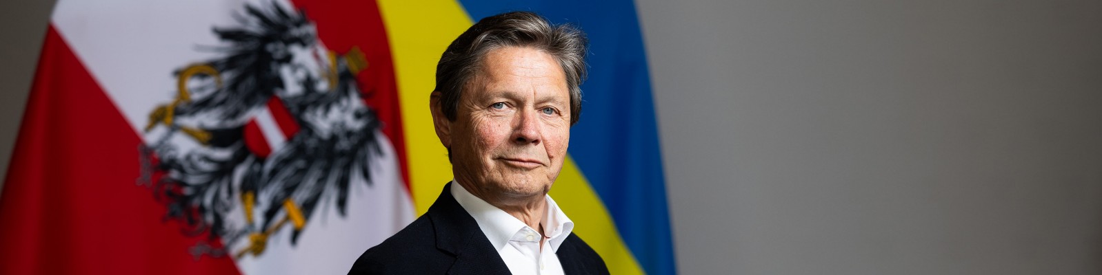 A person in formal attire stands in front of two large flags, one of Austria and one of Ukraine. The setting appears to be official or diplomatic, with a neutral indoor background. The Austrian flag features a prominent black eagle emblem, while the Ukrainian flag displays its characteristic blue and yellow stripes. The overall mood is formal and serious, with a focus on international relations.