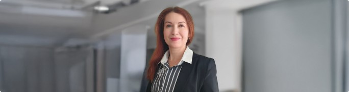 Bogdana Yefremova stands in a modern office hallway. She is dressed in a dark blazer over a striped shirt, suggesting a formal business environment. The background features blurred office architecture with glass partitions and soft lighting. The overall scene conveys professionalism and a contemporary corporate setting.