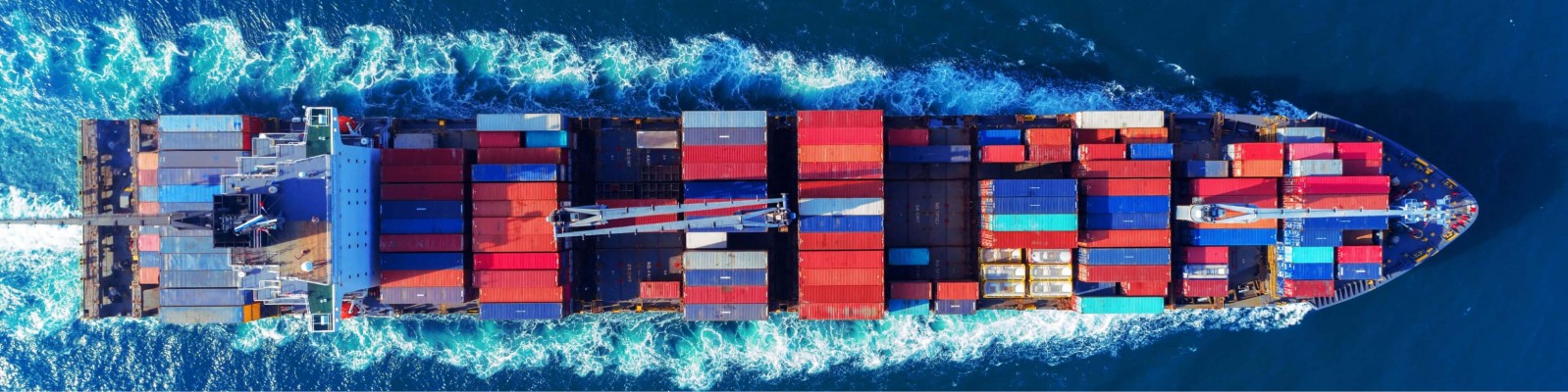 Eine Luftaufnahme eines großen Containerschiffs, das durch den Ozean fährt und mit bunten Versandcontainern beladen ist, vor einem Hintergrund aus lebhaftem blauen Wasser und weißen schaumigen Wellen.