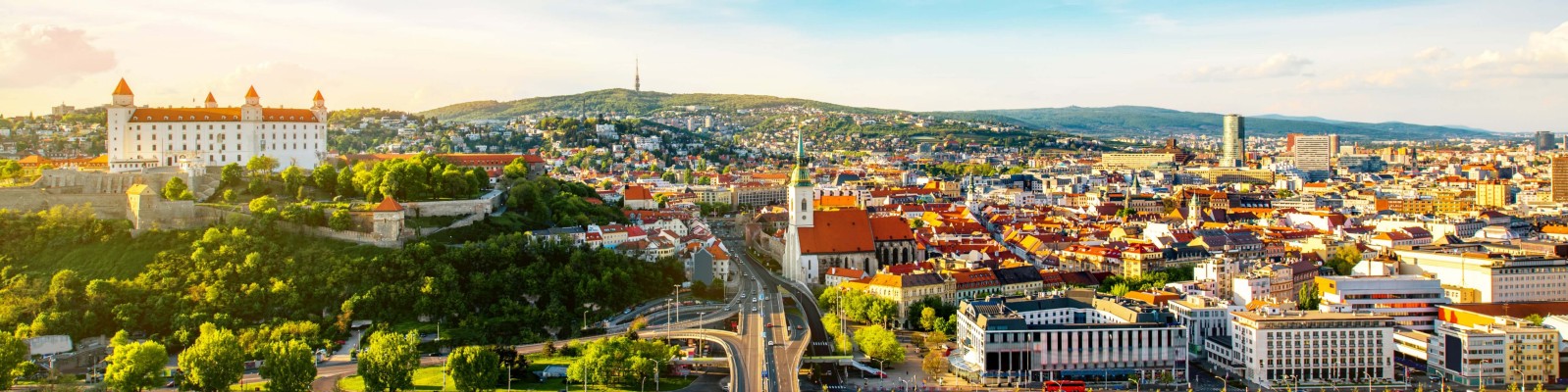A vibrant panoramic view of Bratislava, showcasing the historic Bratislava Castle on a hill surrounded by lush greenery. The cityscape features a mix of modern and traditional architecture under a bright, sunny sky. The image captures the urban layout and scenic beauty of Slovakia's capital city.