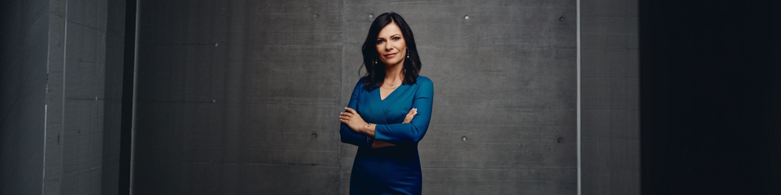 Sladjena Krizaj in a fitted blue dress stands with arms crossed against a dark concrete wall. The setting appears to be an indoor studio or modern industrial space with minimal decor. Her face is blurred, emphasizing anonymity and focusing on body language and posture. The overall composition feels professional and serious, with cool tones and dramatic side lighting.