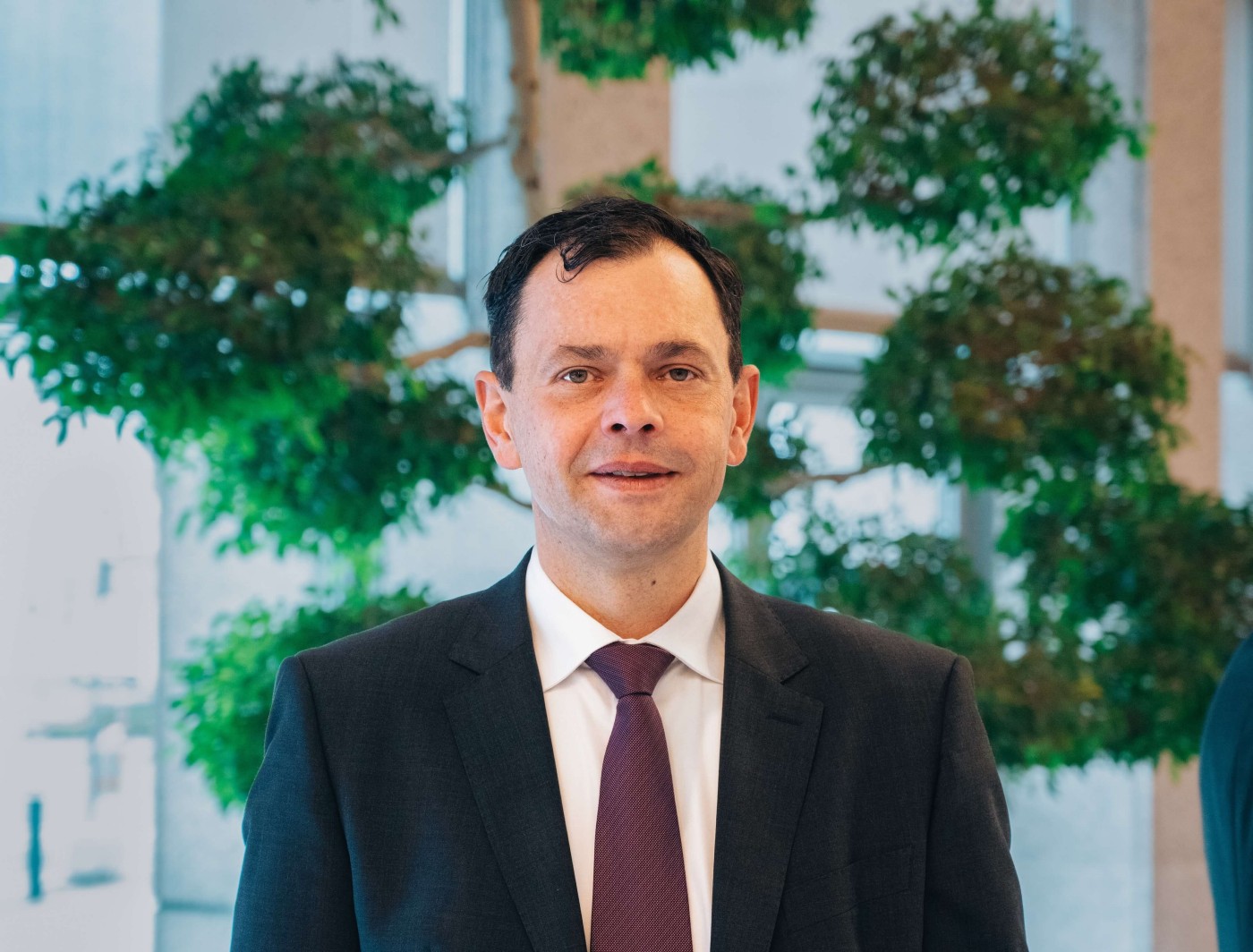 Christian Skopek, gekleidet in einen dunklen Anzug, ein weißes Hemd und eine weinrote Krawatte, steht in einem Innenraum. Im Hintergrund bilden große grüne Pflanzen und natürliches Licht, das durch die Fenster fällt, eine moderne Büro- oder Geschäftsatmosphäre. Die Stimmung ist professionell und geschäftsmäßig, mit Fokus auf formelle Kleidung und einem klaren, zeitgemäßen Ambiente.