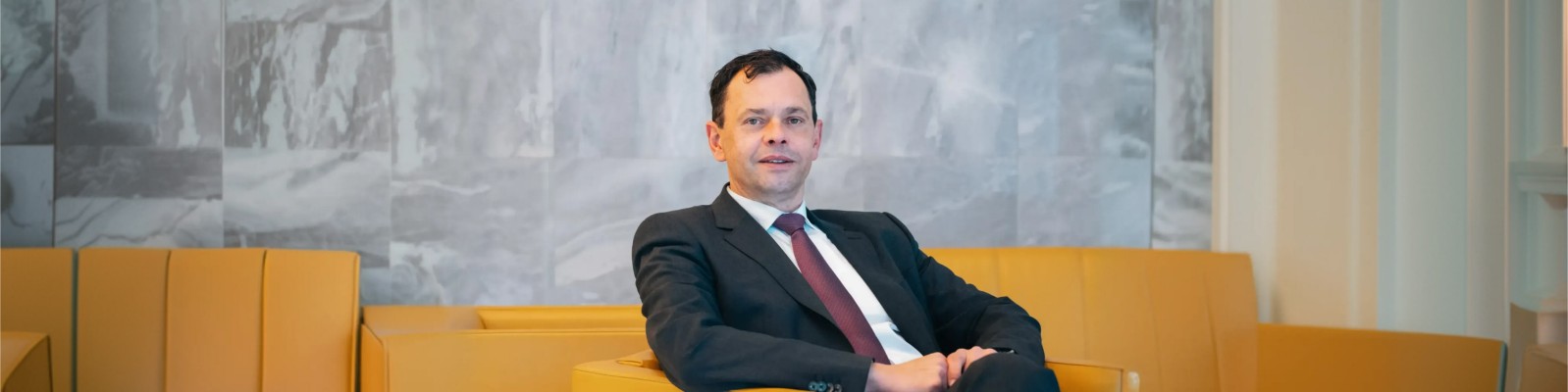 Christian Skopek sitzt in einem dunklen Anzug mit roter Krawatte auf einem modernen gelben Sessel in einem zeitgemäßen Interieur. Im Hintergrund bildet eine Marmor- oder Steinwand in neutralen Farben die Kulisse für eine professionelle und seriöse Atmosphäre. Der Gesamteindruck ist formell und zurückhaltend, klare Linien und gedeckte Farben prägen das Bild.