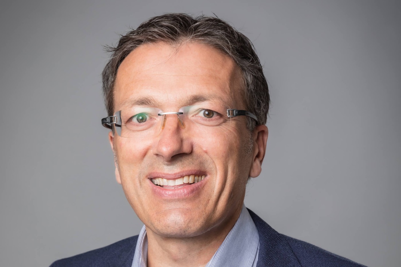 A close-up professional headshot of a business executive with glasses and a friendly smile, representing leadership and confidence.