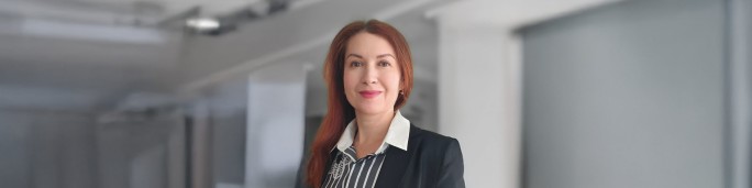 A woman dressed in formal business attire stands in a modern corporate environment. She is wearing a black blazer over a striped shirt, complemented by a white collar. The background features clean architectural lines and soft lighting, suggesting an office or professional workspace.