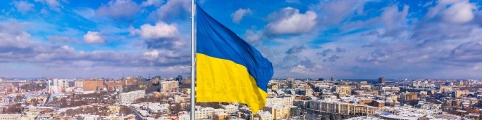 A vibrant Ukrainian flag waves prominently against a backdrop of a sprawling cityscape under a bright blue sky with scattered clouds. The urban setting features snow-covered rooftops and modern architecture, creating a dynamic contrast with the flag's bold yellow and blue colors. The image captures a sense of national pride and a clear winter atmosphere.