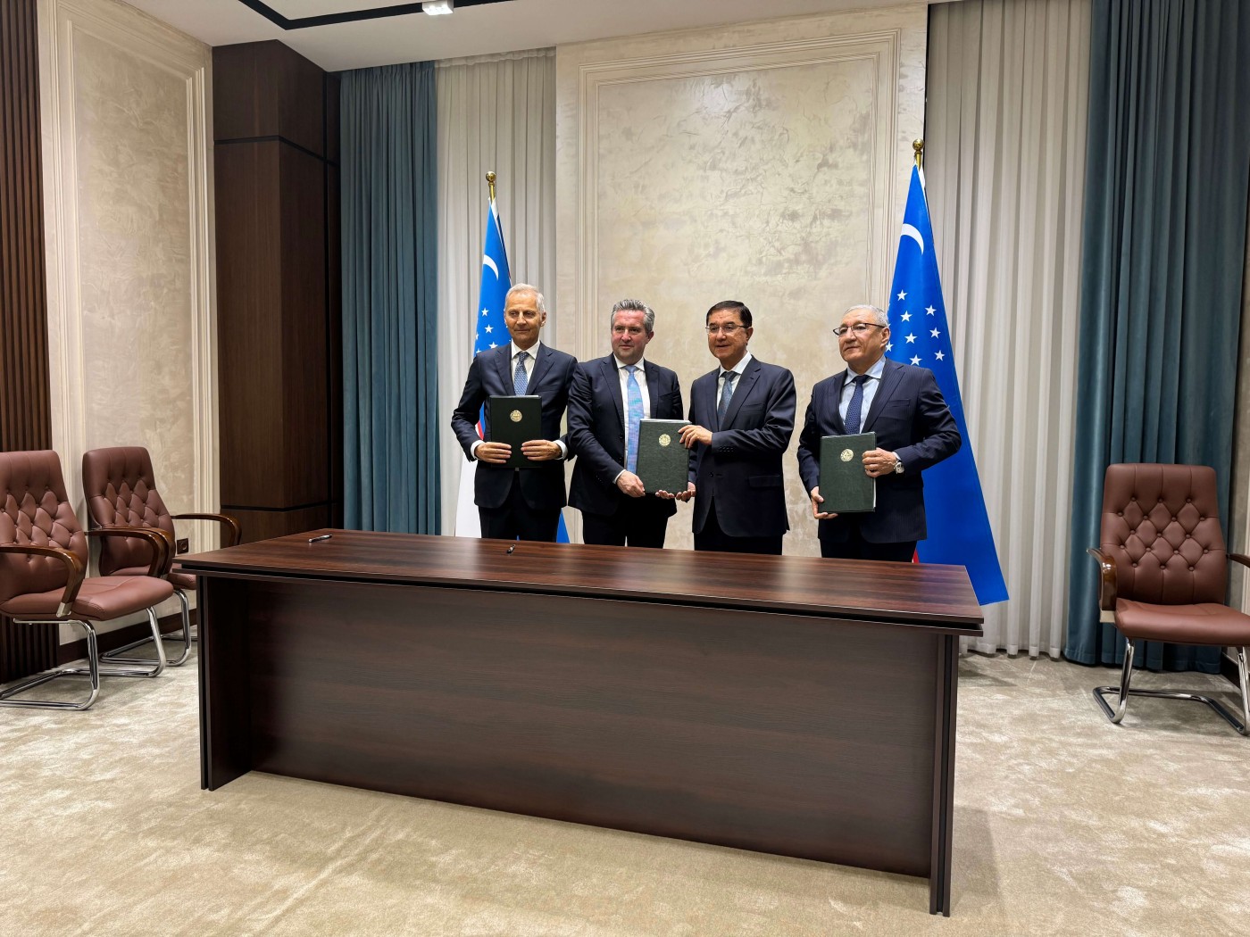Four men in formal business attire stand behind a large wooden conference table holding official-looking folders. The setting appears to be a modern government or corporate meeting room with marble walls, tall curtains, and leather chairs. Two national flags with blue and white colors are displayed behind them, suggesting an official signing or partnership event. The overall atmosphere is professional and ceremonial.
