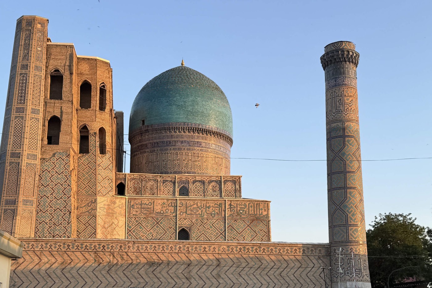 A historic mosque with a large turquoise dome rises against a clear evening sky. The building features intricate geometric tilework and decorative patterns in warm earth tones. A tall, slender minaret stands to the right, also covered in detailed ornamentation. Soft sunset light highlights the textures of the stone and tiles, while the surrounding area remains mostly in shadow.