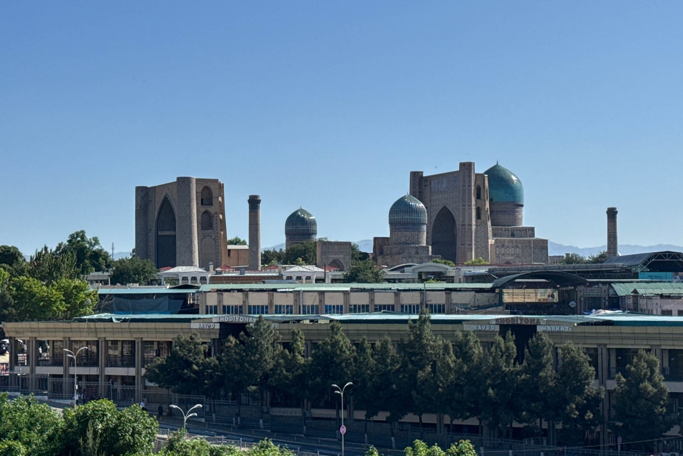 A historic architectural complex with large domes and tall arched structures rises above a modern urban area. The foreground features contemporary buildings and a tree-lined street, contrasting with the ancient stone monuments in the background. Clear blue sky and bright daylight highlight the turquoise domes and geometric forms. The scene blends cultural heritage with everyday city life in a calm, sunny atmosphere.