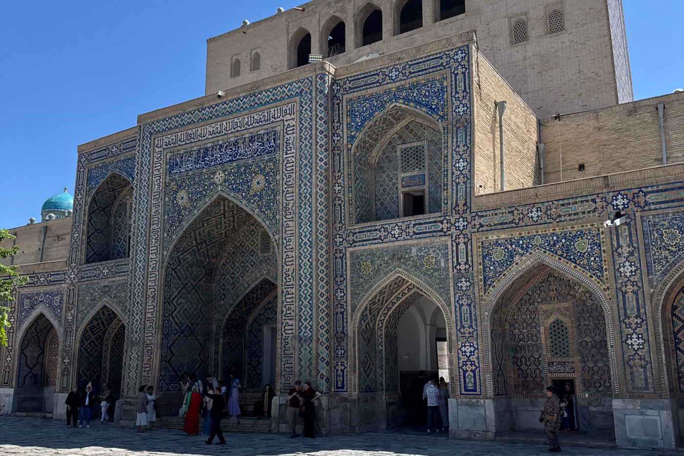 A large historic Islamic building features an ornate facade covered in blue and turquoise geometric tilework. Tall pointed arches frame recessed entrances and windows, creating deep shadows against the sunlit courtyard. People stand and walk near the base of the structure, emphasizing its monumental scale. The architecture suggests a traditional madrasa or mosque in a Central Asian city, with a domed structure visible in the background.