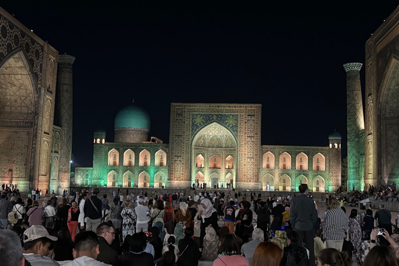 A large crowd gathers at night in front of an illuminated Islamic architectural complex. The central building features a grand arched entrance, intricate tilework, and a turquoise dome glowing against the dark sky. People of various ages stand and sit across the wide courtyard, watching an unseen event. Warm and cool lights highlight the ornate facades and create a festive, atmospheric scene.