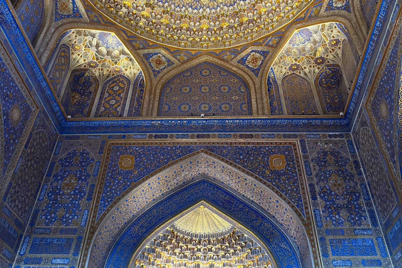 Highly detailed Islamic architectural interior featuring ornate arches and a richly decorated dome. The walls and ceiling are covered with intricate geometric patterns and floral motifs in blue, gold, and white. Light illuminates the gilded details, emphasizing the depth of the muqarnas and tilework. The scene conveys a sense of reverence, craftsmanship, and historic cultural heritage.
