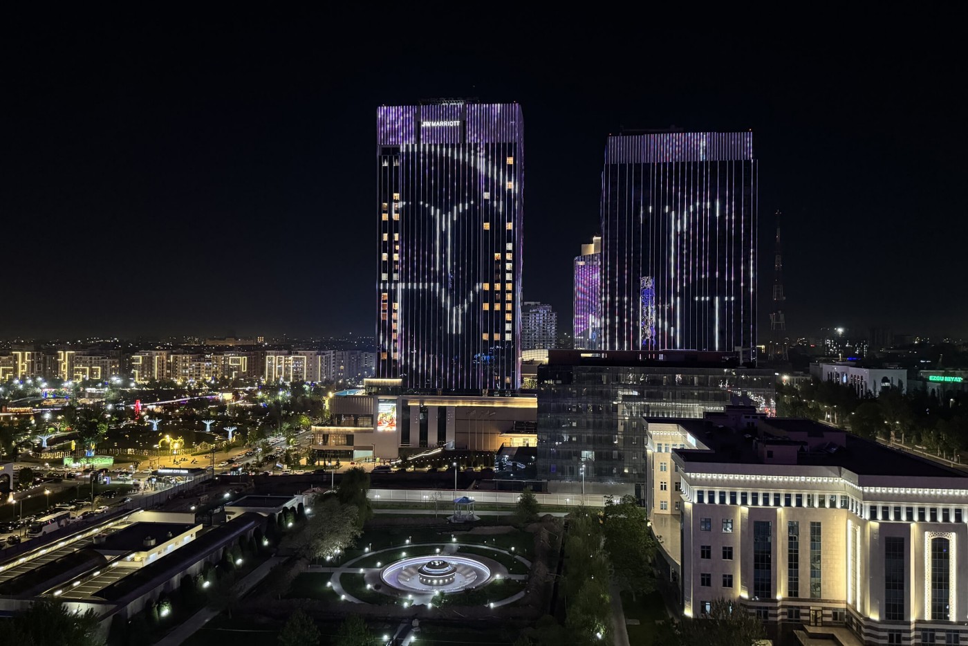 Nighttime cityscape featuring two tall modern glass towers with animated light displays on their facades. The buildings overlook a well-lit urban area with streets, smaller structures, and distant lights stretching toward the horizon. In the foreground, a circular fountain or plaza with decorative lighting sits within a landscaped park. The scene highlights contemporary architecture, vibrant city life, and dramatic contrast between bright lights and dark sky.