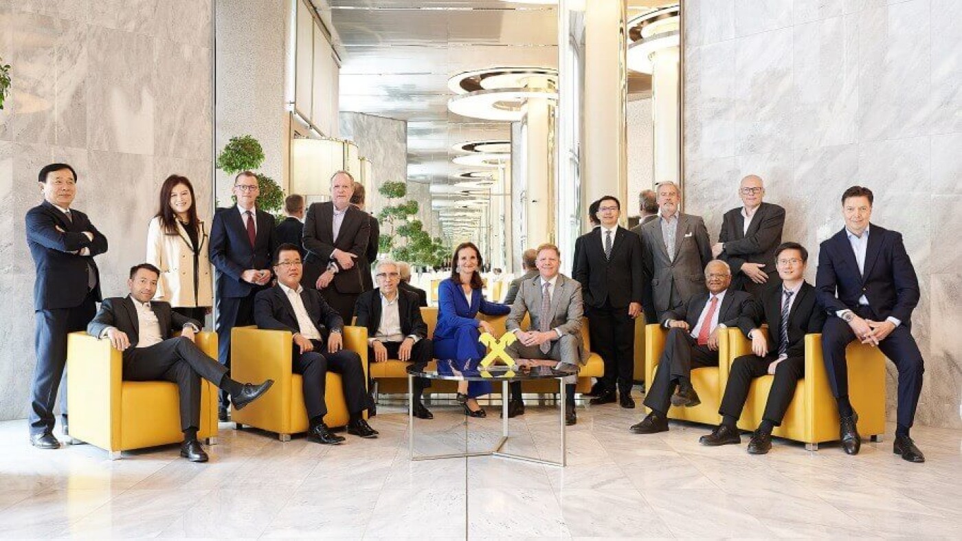 A diverse group of professionals seated and standing in a luxurious lounge area with marble walls and yellow chairs. The setting features modern decor, reflective surfaces, and indoor greenery. The individuals are dressed in formal attire, showcasing a mix of genders and ages.