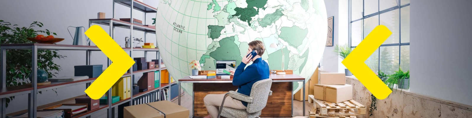 A man sits at a desk in a modern office setting, talking on the phone. The backdrop features a large green world map mural, adding a global theme to the space. Shelves with books and boxes are visible, suggesting an organized workspace.