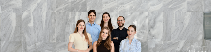A group of six young adults stands together in front of a large gray marble wall. They are dressed in smart casual and business attire, suggesting a professional or corporate setting. The composition is centered on the people, with neutral stone textures filling the background. The overall mood is friendly, confident, and contemporary.