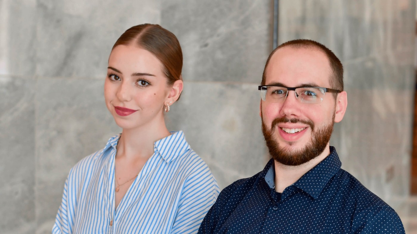 Two young professionals pose together in a modern indoor setting. The woman wears a light blue striped shirt, while the man wears glasses and a dark blue patterned shirt. They both smile confidently toward the camera, suggesting a friendly and approachable work environment. The neutral gray background keeps the focus on their expressions and professional appearance.