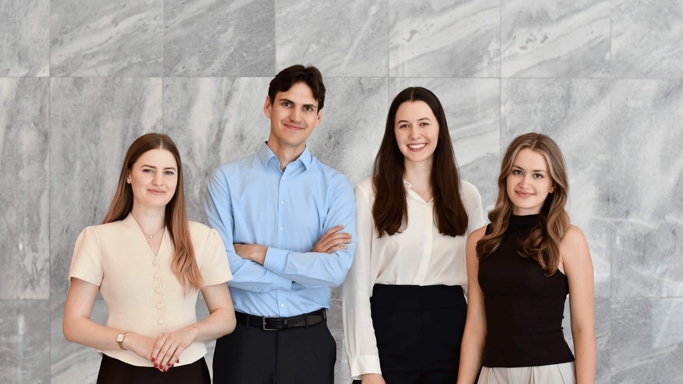 A group of four young professionals stands side by side against a light gray marble wall. They are dressed in smart casual office attire, including blouses, a button-down shirt, and skirts. The individuals smile confidently, suggesting a friendly and collaborative work environment. The neutral stone background keeps the focus on the people and their professional appearance.