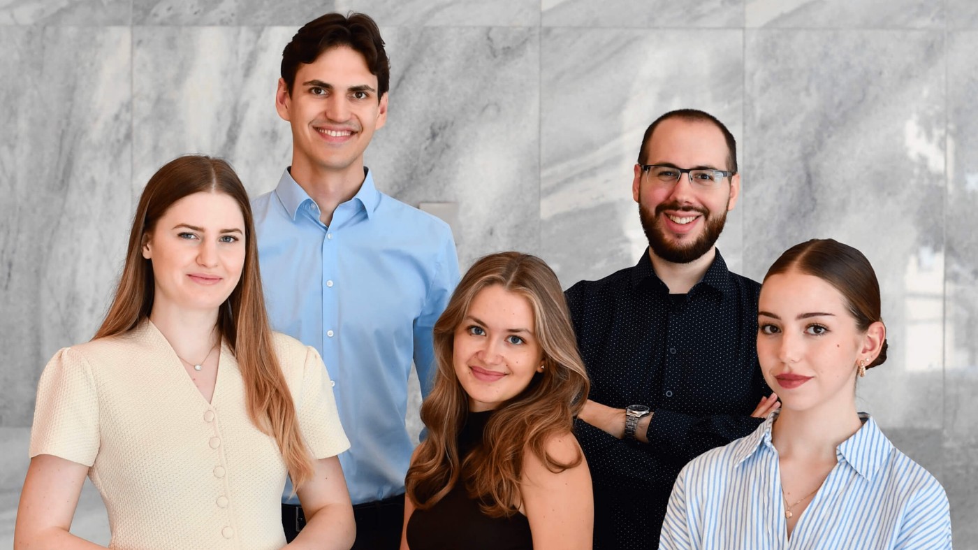 A group of five young professionals stands together in front of a light marble wall. They are dressed in smart casual and business attire, including button-up shirts and a light dress. The group smiles confidently at the camera, suggesting a friendly and collaborative team atmosphere. The neutral stone background keeps the focus on the people and their expressions.