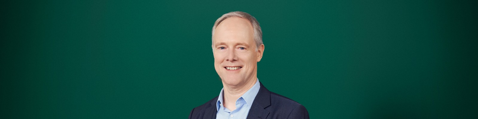 Porträt eines Mannes mittleren Alters in Businesskleidung vor einem einfarbig grünen Hintergrund. Er trägt ein dunkles Sakko über einem hellblauen Hemd und lächelt freundlich in die Kamera. Das Bild ist klar und minimalistisch gestaltet und konzentriert sich auf Gesicht und Oberkörper des Mannes. Der schlichte Hintergrund unterstreicht seine professionelle und sympathische Ausstrahlung, die sich für den Einsatz im Unternehmensumfeld eignet.