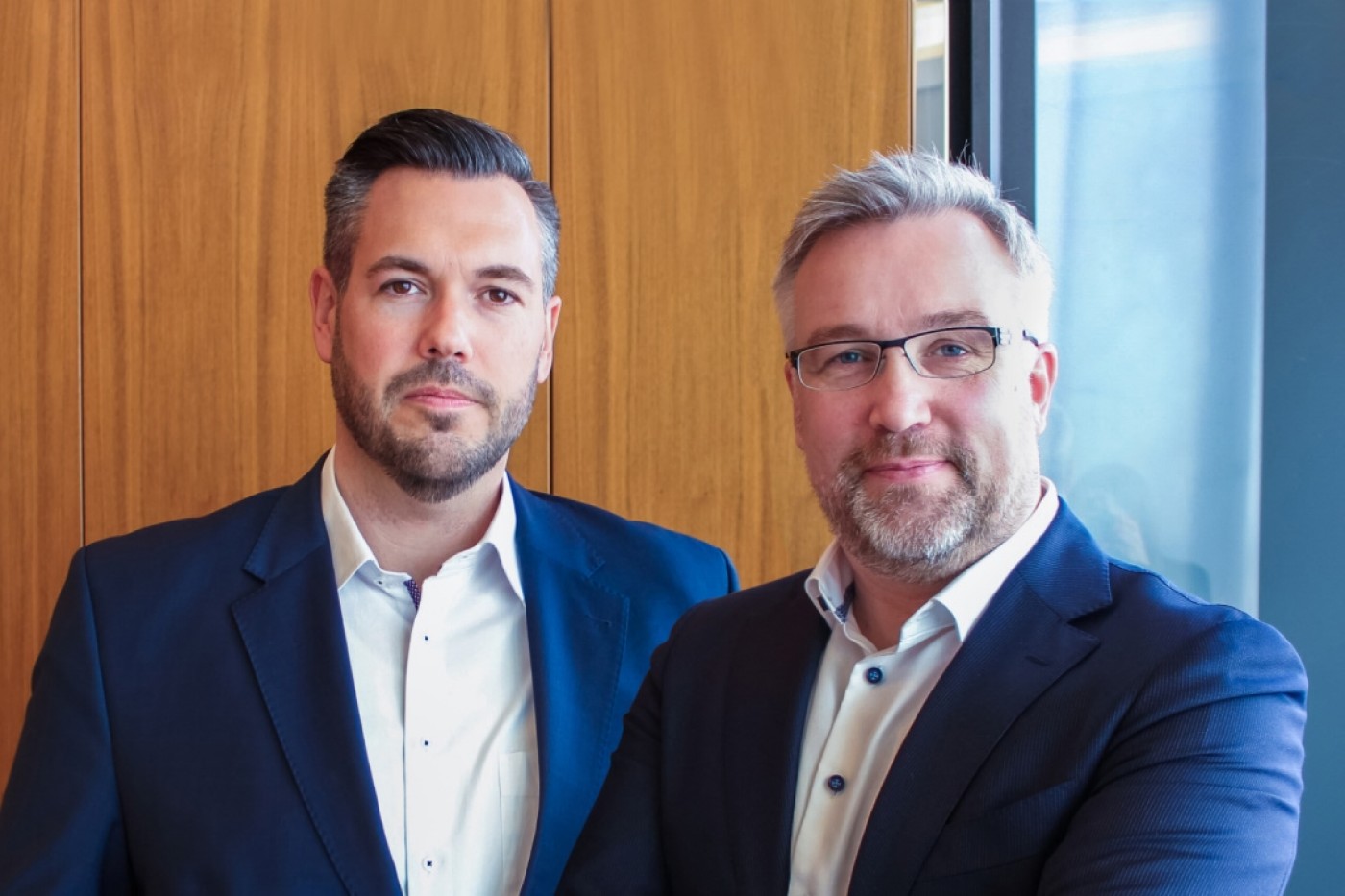 Two professional men in business attire standing in front of a wooden background, representing leadership in business consulting.