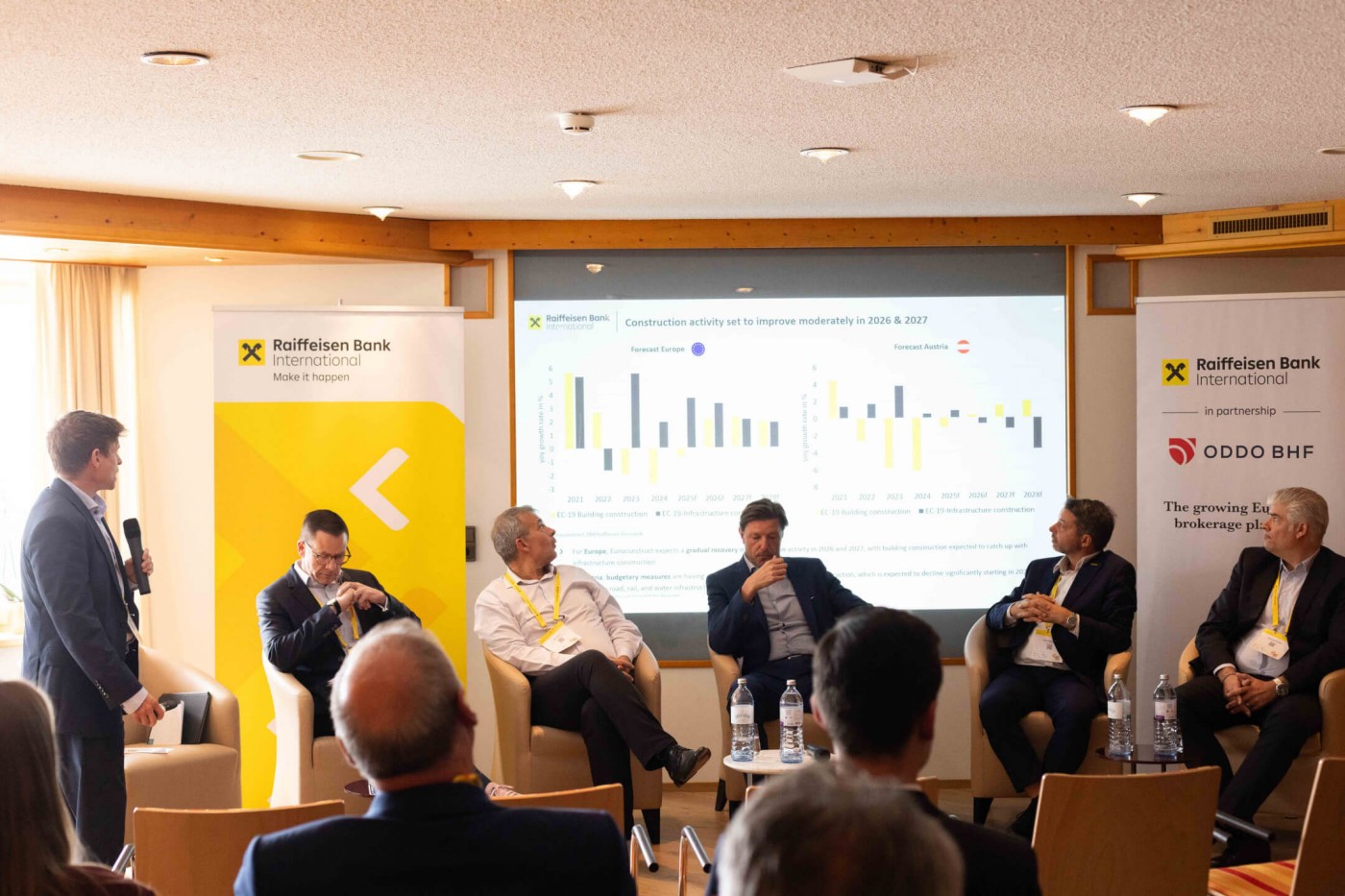 Eine Gruppe von Geschäftsleuten nimmt an einer Podiumsdiskussion in einem Konferenzraum teil. Ein Mann steht mit einem Mikrofon, während fünf weitere in Sesseln vor einer großen Projektion sitzen. Die Präsentationsfolie zeigt Balkendiagramme, die Daten aus den Jahren 2016 und 2017 vergleichen. Die Balken sind gelb und blau und mit verschiedenen Beschriftungen versehen. Banner von Finanzinstituten sind zu beiden Seiten der Leinwand angebracht und unterstreichen die seriöse Atmosphäre. Die Stimmung ist formell und konzentriert, die Beleuchtung angenehm warm.
