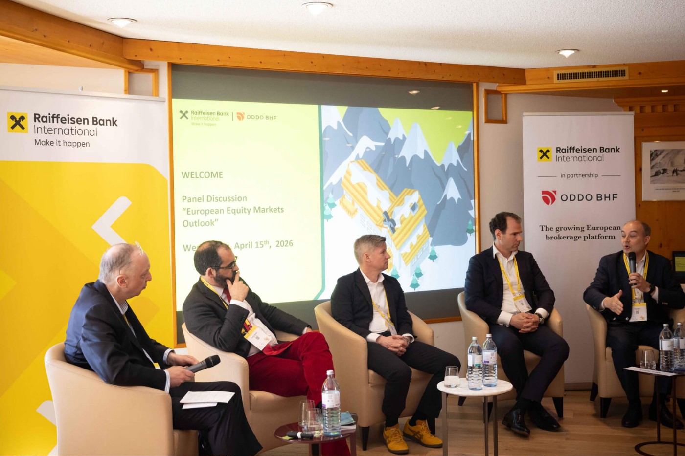 Vier Geschäftsleute sitzen auf einer Bühne und nehmen an einer Podiumsdiskussion teil. Im Hintergrund ist eine Präsentationsfolie mit dem Titel „Ausblick auf die europäischen Aktienmärkte“ und dem Datum Mittwoch, 10. April 2019, zu sehen. Zwei vertikale Banner der Raiffeisen Bank International und eines weiteren Finanzinstituts flankieren das Podium. Der Raum wirkt wie ein Konferenzraum mit warmen Holzwänden und einem kleinen Publikumsbereich davor. Wasserflaschen und Mikrofone auf kleinen Tischen unterstreichen die professionelle Konferenzatmosphäre.