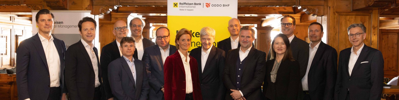 Eine Gruppe von Geschäftsleuten posiert für ein formelles Gruppenfoto bei einer Firmenveranstaltung in geschlossenen Räumen. Sie tragen Businesskleidung, darunter Anzüge und Blazer, und stehen vor einem leuchtend gelben Banner. Der Raum wirkt holzgetäfelt, möglicherweise ein Konferenz- oder Besprechungsraum, und ist warm beleuchtet. Die Atmosphäre ist professionell und kooperativ und deutet auf ein Geschäftstreffen, die Bekanntgabe einer Partnerschaft oder eine Netzwerkveranstaltung hin.
