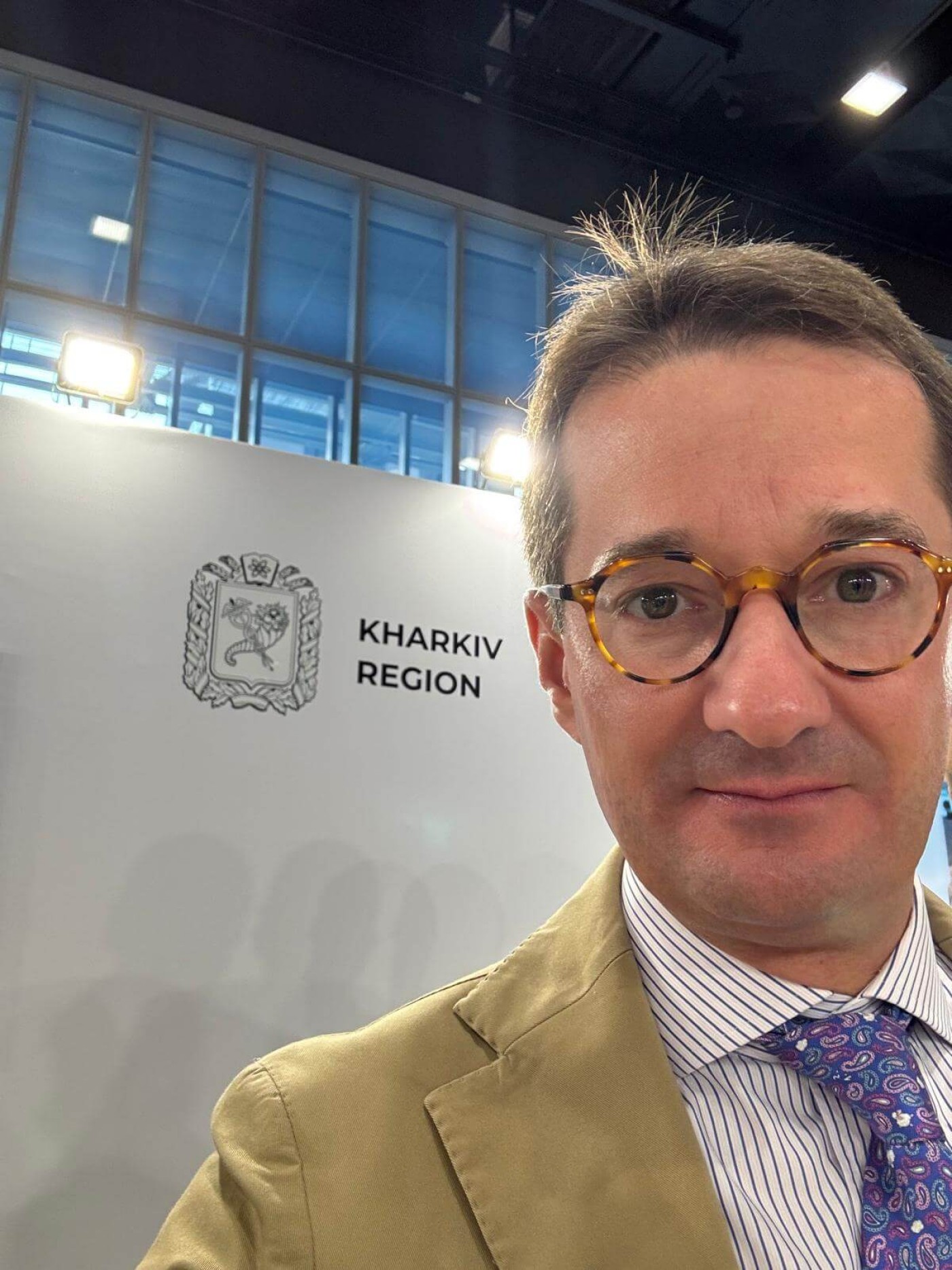 A person in formal attire stands in front of a backdrop featuring the Kharkiv Region emblem and text. The setting appears to be indoors, with bright lighting and large windows visible in the background. The image highlights the official branding and professional environment.