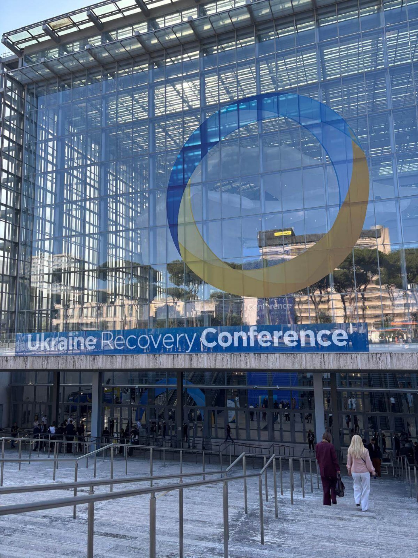 A large glass-fronted building hosts the Ukraine Recovery Conference, with visible signage featuring the event name. The setting includes a modern architectural design with reflective glass panels and a spacious entrance. Two individuals are seen walking toward the venue, adding a human element to the scene.