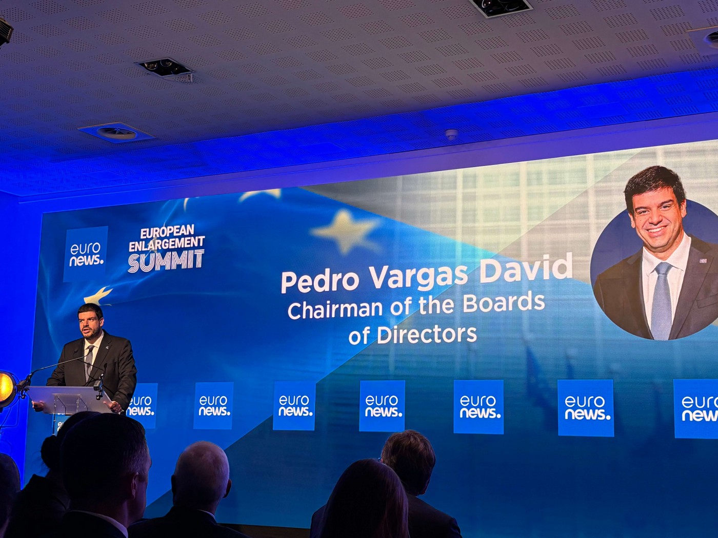 A suited speaker stands at a podium addressing an audience during a formal conference. Behind him, a large illuminated screen displays the text "EUROPEAN ENLARGEMENT SUMMIT" and "Pedro Vargas David Chairman of the Boards of Directors" alongside a portrait of a man in a suit. The stage is lit with blue lighting and features multiple "euro news." panels. Silhouetted attendees are visible in the foreground, emphasizing the professional setting.