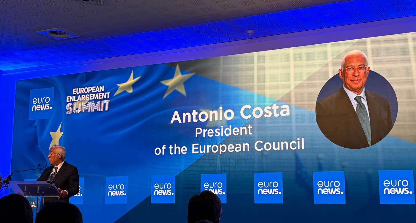 A formal conference stage is set for the European Enlargement Summit. A speaker stands at a podium on the left, addressing the audience in front of a large illuminated backdrop. The backdrop displays the text "EUROPEAN ENLARGEMENT SUMMIT" and "Antonio Costa President of the European Council" alongside a blurred portrait. Multiple blue screens with the "euro news." text are visible across the stage. The overall atmosphere is professional and political, with blue lighting and European flag imagery.