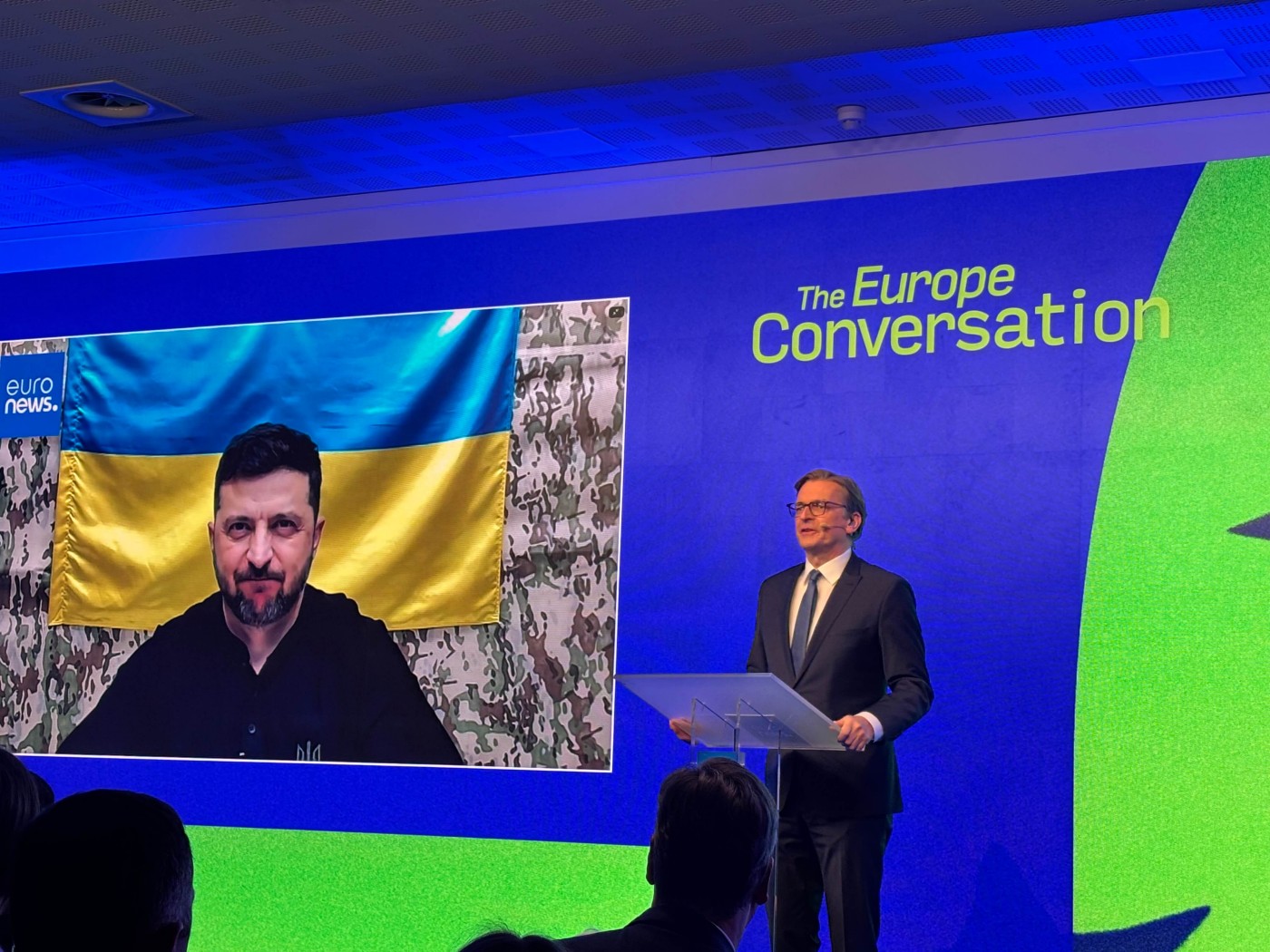 A formal conference scene shows a suited speaker standing at a clear podium on a stage. Behind him, a large screen displays a person speaking remotely in front of a Ukrainian flag and camouflage backdrop. The stage backdrop reads "The Europe Conversation" with bright blue and green lighting. The audience is visible in silhouette, emphasizing the event’s public and political nature. The overall atmosphere is serious and focused on European and Ukrainian topics.