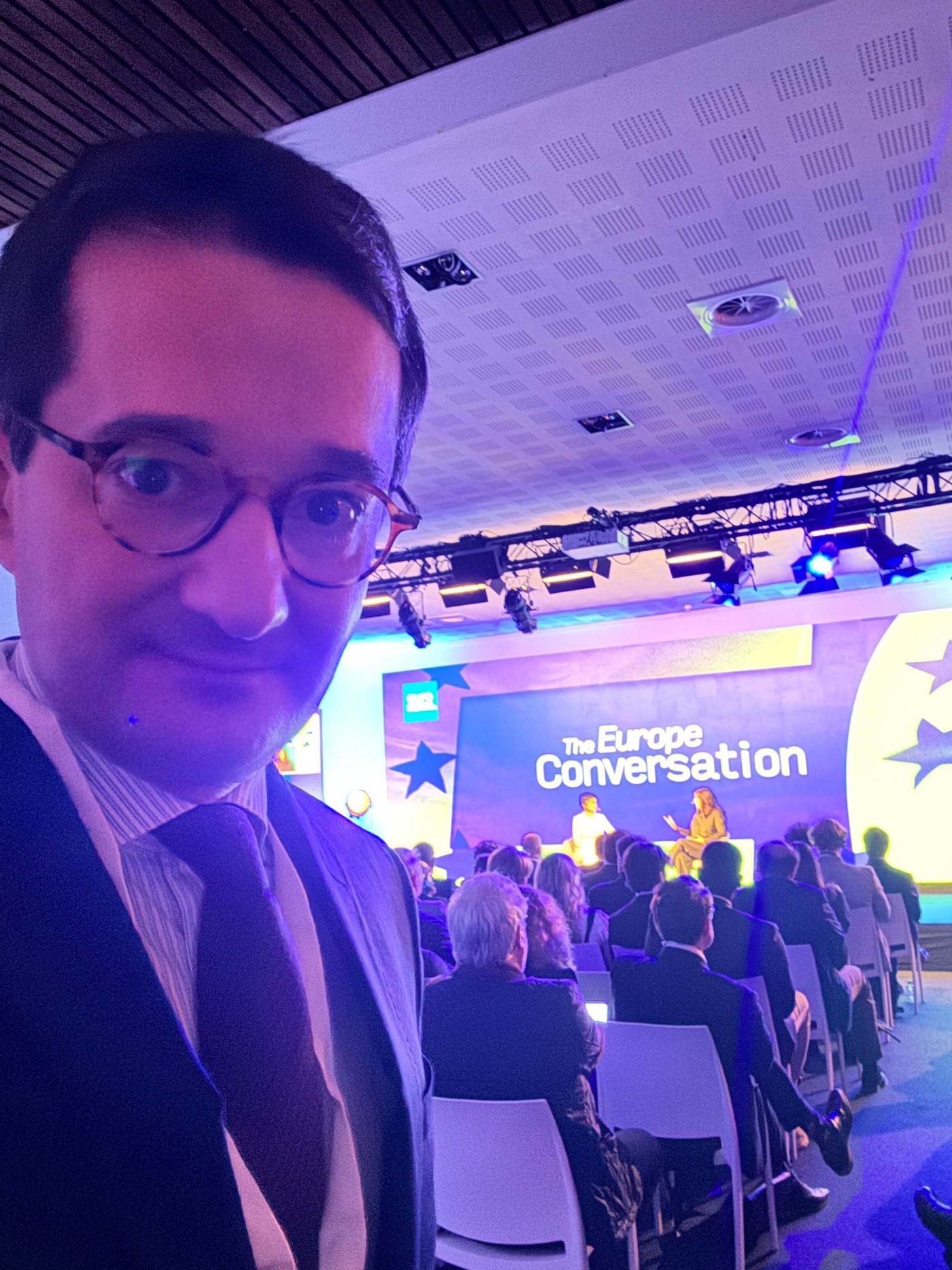 Paul Pasquali formally dressed, takes a selfie in front of a conference stage. The stage backdrop prominently displays the text "The Europe Conversation" with large star graphics. Rows of seated participants face the brightly lit speakers on stage in a modern auditorium. Cool blue and purple lighting creates a professional yet dynamic atmosphere. The scene suggests a high-profile discussion or panel focused on European topics.