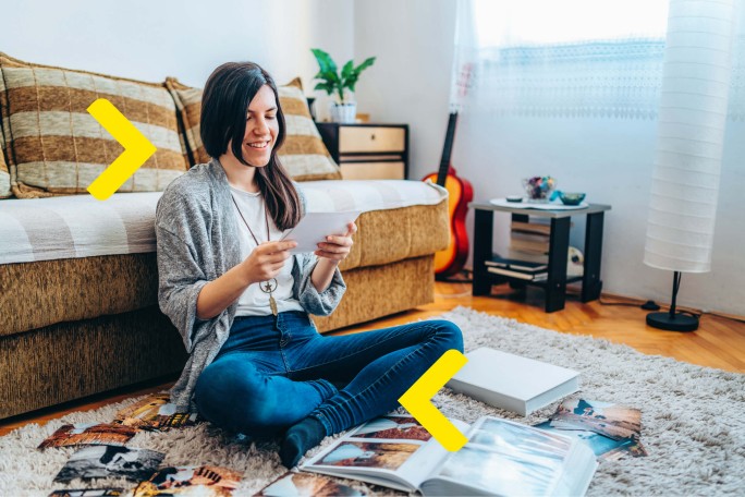 A woman sitting cross-legged on a rug, reading a magazine with photos, in a well-lit living room with a sofa, guitar, and plants.