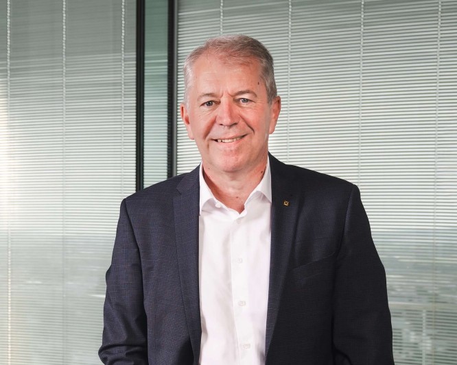 Erwin Tinhof, in dunklem Anzug und hellem Hemd, lächelt in einem modernen Büro. Er steht vor großen Jalousien und einem Glasfenster, was auf ein Geschäftsumfeld mit Blick auf die Stadt hindeutet. Die Szene vermittelt Professionalität, Selbstbewusstsein und Zugänglichkeit. Neutrale Farben und sanftes Licht schaffen eine klare, geschäftsmäßige Atmosphäre.