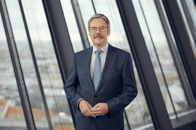 Erwin Hameseder in a dark blue suit stands confidently in front of tall glass windows. He wears a light blue tie and glasses, with his hands gently clasped. The background shows a blurred cityscape seen through the large window panels. Soft natural light creates a professional and corporate atmosphere.