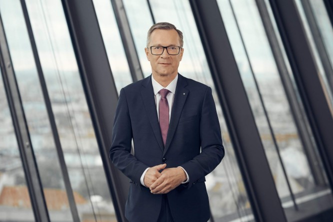 Heinz Konrad stands in a modern office environment. He is dressed in a dark suit, white shirt, and a solid-colored tie, looking directly at the camera with a composed expression. Large floor-to-ceiling windows behind him reveal an out-of-focus urban landscape. The overall scene conveys professionalism and corporate leadership.