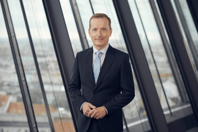 Manfred Wilhelmer  in a dark suit and light blue tie stands confidently in a modern office environment. He faces the camera with a slight smile and his hands gently clasped. Tall floor-to-ceiling windows behind him reveal an urban cityscape in soft focus. The overall color palette is neutral and professional, emphasizing a corporate and executive atmosphere.
