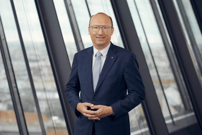Reinhard Schwendtbauer  in a dark blue suit poses for a professional portrait. He stands in front of large floor-to-ceiling windows in a modern high-rise office. The cityscape is softly visible in the background, creating a corporate atmosphere. The lighting is soft and even, emphasizing a formal and confident business mood.