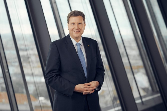 Thomas Wass in a dark suit and light blue tie stands confidently in a modern office environment. He smiles at the camera with his hands gently clasped in front of him. Large floor-to-ceiling windows behind him reveal an out-of-focus cityscape. The overall scene conveys professionalism and a corporate atmosphere.