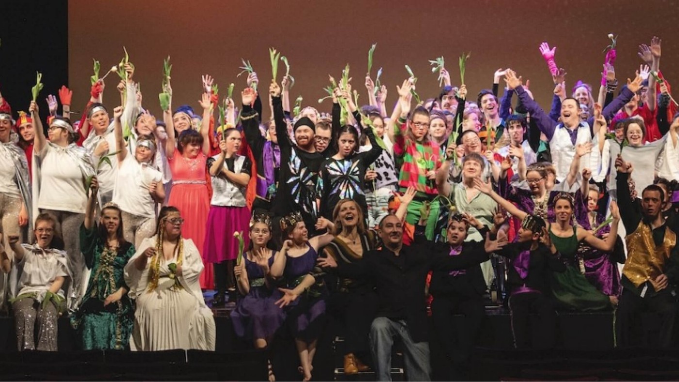 Eine große Gruppe von Künstler:innen in farbenfrohen Kostümen feiert auf der Bühne, hält Blumen und reckt die Hände in die Höhe.