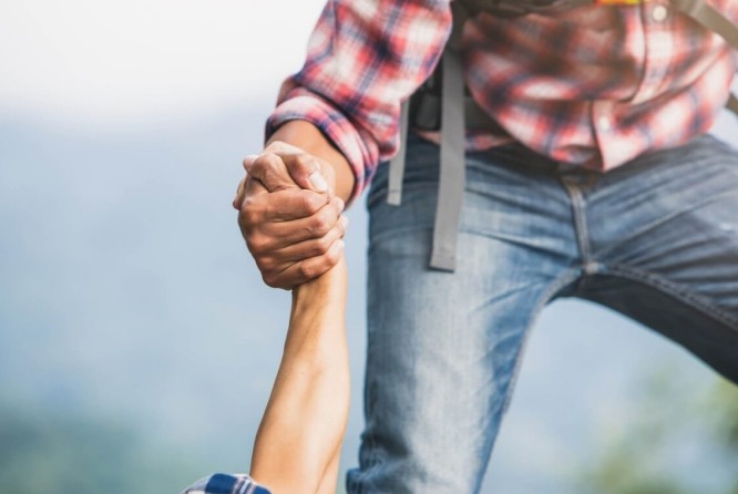 Eine Person in einem karierten Hemd und Jeans reicht jemandem beim Bergsteigen die Hand, um ihm zu helfen.