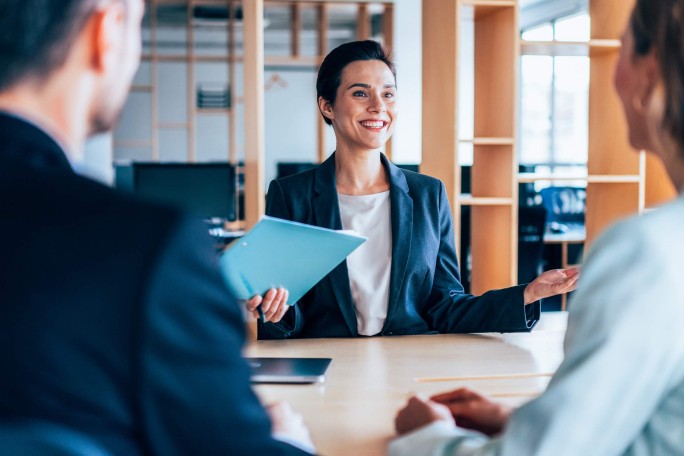 Dieses Bild zeigt eine professionelle Geschäftssituation, in der eine Person mit einer Mappe Informationen präsentiert und mit zwei Kolleg:innen interagiert. Die Umgebung spiegelt eine kooperative Atmosphäre wider und betont Kommunikation und Teamarbeit im Unternehmenskontext.