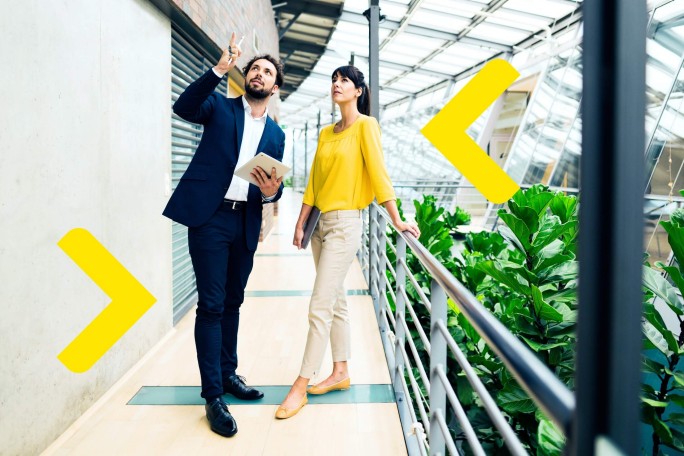 Zwei Geschäftsleute, ein Mann und eine Frau, führen ein Gespräch in einem hellen, modernen Innenraum mit Pflanzen und Glasarchitektur. Der Mann hält ein Tablet und gestikuliert, während die Frau an einem Geländer lehnt und einen Laptop trägt.